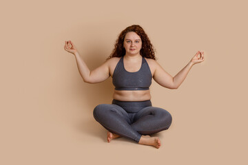 Curly haired calm tranquil joyful barefoot woman sitting in lotus ashtanga asana yoga position, crossing legs, meditating. Wearing top and sportive gray sportswear. Problems with bones, spin treatment