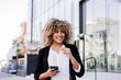 © Eva - beautiful smiling business woman using mobile phone and headphones in city. Buildings background