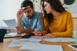 © Tijana - Serious Couple Paying Bills Together at Home. Worried man and woman talking about family budget and savings, calculating household bills and expenses while sitting at table with laptop and coffee.