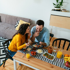 Wall Mural - High angle view of smiling married couple looking at phone and laughing while having breakfast together at home