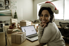 Young Woman In Santa Hat Using Laptop Next To Christmas Gifts In Studio