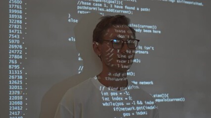 Chest up slowmo of young Caucasian man in white tee and eyeglasses standing against projector with running program code on dark background projected over his face