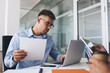 © BullRun - Professional male employer in optical eyeglasses analyzing paper report while browsing financial website on modern laptop computer, Caucasian businessman in spectacles reading publication on netbook