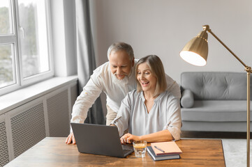 Old man and an age woman sit at home at a table in a laptop. Charming woman and gray-haired man with a beard surf on the Internet. Emotions of surprise and joy.