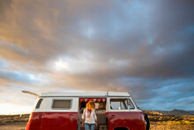 People Travel And Enjoy Vanlife Adventure Lifestyle. Standing Woman Against A Red Classic Vintage Van. Beautiful Sunset Colors Sky In Background. Concept Of Camper Holiday Vacation