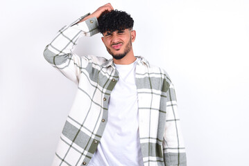 young arab man with curly hair wearing overshirt over white background saying: Oops, what did I do? Holding hand on head with frightened and regret expression.