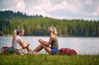 © luckybusiness - Two woman sitting in grass in front of lake and talking with backpacks by their side. Leirsure time. Trip, weekend, fun, lifestyle, nature concept