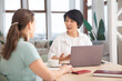 © ulza - Happy multiethnic colleagues sitting together at coworking place, more experienced employee shares. The adorable Asian businesswoman advises the client in the office