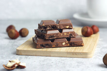 A Bar Of Milk Chocolate With Nuts And Cookies Lies On A Wooden Board On A Gray Table. Hazelnuts Are Scattered Behind And There Is A Mug Of Tea. The Concept Of Natural Chocolate Without Palm Oil. 