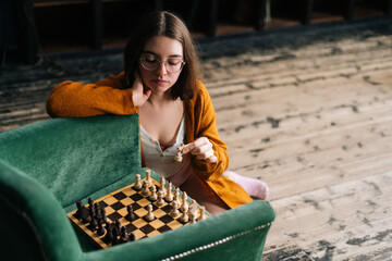 Wall Mural - High-angle view of attractive young woman wearing elegant eyeglasses making chess move with knight piece sitting in wooden floor. Pretty intelligent lady playing logical board game alone at home.