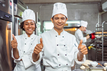 Young  African  female and  senior asian male bakers looking at camera..Chefs  baker in a chef dress and hat, cooking together in kitchen.Team of professional cooks in uniform.