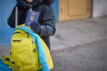 Evacuation Of Civilians, Sad Child With The Flag Of Ukraine. Refugee Family From Ukraine Crossing The Border. Hand Holding A Passport Above The Luggage With Yellow-blue Flag. Stop War, Support Ukraine