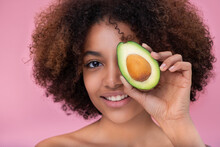 Portrait Of A Beautiful Young Dark Skinned Woman With Curly Hair Covers One Eye With Half An Avocado Looks At The Camera And Smiles On A Pink Background.