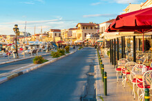 The Main Street Running Through The Picturesque Village Of Aegina, On The Island Of Aegina Greece, With Shops And Cafes On One Side And The Marina And Port Across.