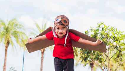 Happy Asian funny child or kid little boy smile wear pilot hat and goggles play toy cardboard airplane wing flying against summer sky cloud on trees garden background, Startup freedom concept