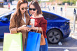 © Tom Wang - Happy  young woman standing on city parking near electric car, charging automobile battery from small city station, holding shopping bags and watching smart phone