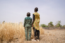 Rear View Of Two African Boys Standing In Front Of A Barren Field With Tall Dry Grass Staring At The Horizon, Questioning Their Fate; Global Warming, Desertification And Water Scarcity Concepts
