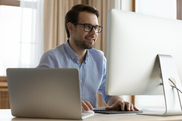 Smiling young man software developer sit at table work on two computer devices write program code. Confident male designer look on pc screen create project at modern office using both laptop desktop