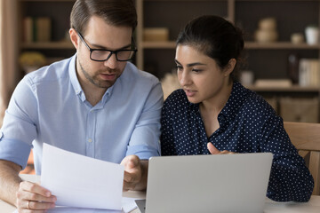 Serious multiethnic business partners coworkers students Indian woman Caucasian man sit at desk discuss document read sales report. Two young diverse teammates engaged in paperwork solve problem talk