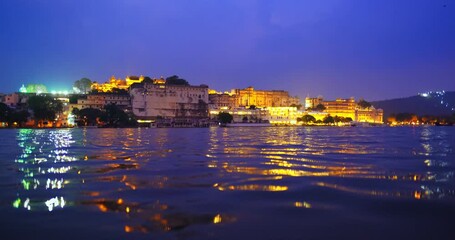 Fototapete - Udaipur City Palace on the bank of beautiful lake Pichola at sunset - Rajput architecture of Mewar dynasty rulers of Rajasthan. Water ripples reflect night city view. Udaipur, India