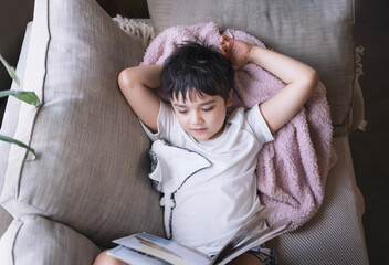 Wall Mural - Happy School kid lying on sofa reading a book with morning light shining from window. Top view Child boy reading story relaxing at home on weekend, Positive children concept