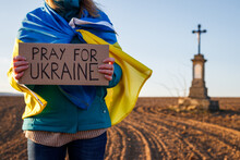 Pray for Ukraine. Woman wrapped in ukrainian flag holding anti war sign cardboard outdoors. Defocused religious cross  monument
