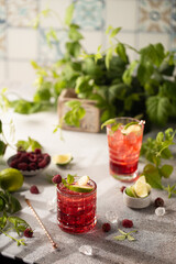 Wall Mural - Raspberry cocktail with lime,mint,ice cubes and fresh berries on grey background with juicy summer greenery.Beautiful photo of refreshing drink.