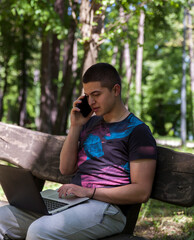 A young handsome man is sitting on a wooden bench and working on his laptop in nature.