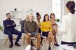 © Studio Romantic - Female business coach in meeting in office speaks in front of small group of business people. Group of multiracial diverse contented people sitting on chairs attentively listening to their leader.