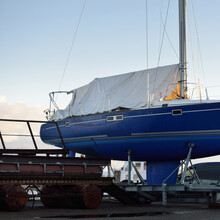 Blue Sloop Rigged Yacht Standing On Land In A Small Harbor On A Clear Winter Day. Waiting For The New Sailing Season. Industry, Business, Transportation, Service, Logistics, Sport, Recreation Concepts