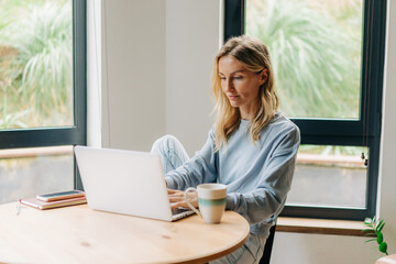 A young woman in home clothes sits at a table at home and works hard at a laptop at a remote job