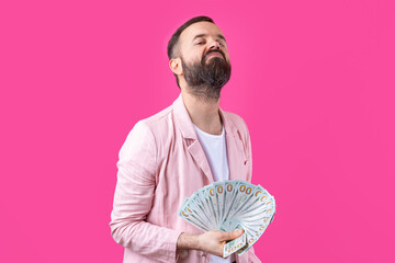 Portrait of a contented young businessman with a beard dressed in a pink jacket showing us dollar banknotes against a red studio background. Taste, smell of money.