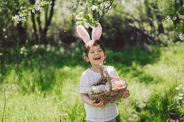 Easter egg hunt in spring garden. Funny boy with eggs basket and bunny ears on Easter egg hunt in sunny spring garden. Children celebrating Easter. Happy easter card