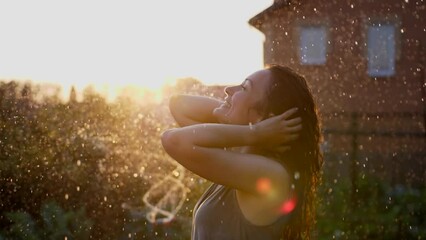 Wall Mural - Happy smiling woman is under the rain. Stunning golden sun rays shine on playful young woman enjoying a summer rain. slow motion