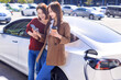 © Tom Wang - Happy  young woman standing on city parking near electric car, charging automobile battery from small city station, watching smart phone and checking information