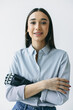 Vertical image of businesswoman in striped shirt keeping arms crossed, smiling at camera against white wall, having bionic prosthesis instead of left hand after terrible trauma and injury
