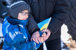 © sokorevaphoto - Families against war. Upset Ukrainian boy with his grandmom, with bue yellow flag, protesting war conflict. Evacuation of civilians. People on the background of destruction