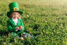 St. Patrick's Day. A Girl In A Green Suit Is Sitting In A Large Clearing Of Clover, Holding A Pot With Gold Coins In Her Hands. Copyspase. Happy Joyful Toddler Baby