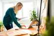 © baranq - Young woman standing by a desk using laptop holding documents working from home office