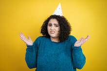 Young Beautiful Woman Wearing A Birthday Hat Over Isolated Yellow Background Clueless And Confused Expression With Arms And Hands Raised