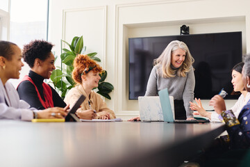 Portrait of senior white businesswoman with laptop smiling and listening to multiethnic female colleagues in boardroom