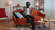 © Andrii Iemelianenko - African American man wearing VR headset resting at home sitting on sofa in living room alone using futuristic technology making gestures with hands in air, play 3D video game. Virtual reality glasses