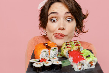 Close up hungry starving young woman in casual clothes hold in hand look at makizushi sushi roll served on black plate traditional japanese food show tongue isolated on plain pastel pink background