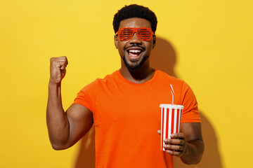 Wall Mural - Young smiling fashionable fun happy man of African American ethnicity 20s wear orange t-shirt stylish red glasses hold cup of soda pop fizzy water isolated on plain yellow background studio portrait