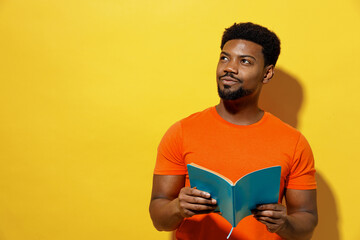 Wall Mural - Young minded fun student man of African American ethnicity 20s wear orange t-shirt read novel book learn look aside dream isolated on plain yellow background studio portrait. People lifestyle concept.