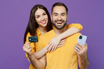 Wall Mural - Young happy couple two friends family man woman together in yellow clothes use mobile cell phone hold credit bank card shopping online order delivery booking tour isolated on plain violet background