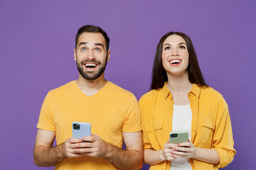 Wall Mural - Young smiling happy fun couple two friends family man woman together in yellow casual clothes hold use mobile cell phone look overhead on workspace isolated on plain violet background studio portrait.