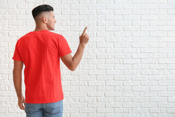 Wall Mural - Handsome young man in stylish t-shirt pointing at something on white brick background, back view