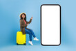 Happy young black woman sitting on suitcase and pointing at smartphone with mockup for online travel agency website