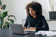 © Kateryna - Young adult African American business woman in the office. Attractive confident smart woman top manager or trader, sits in front of a laptop, looks the screen, makes working notes in a notebook